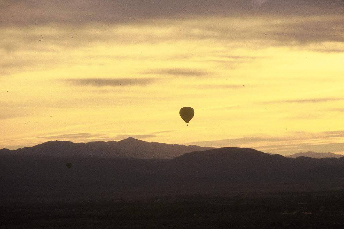 A lone hot air balloon floats above dark mountain ridges beneath a layered golden sunset.