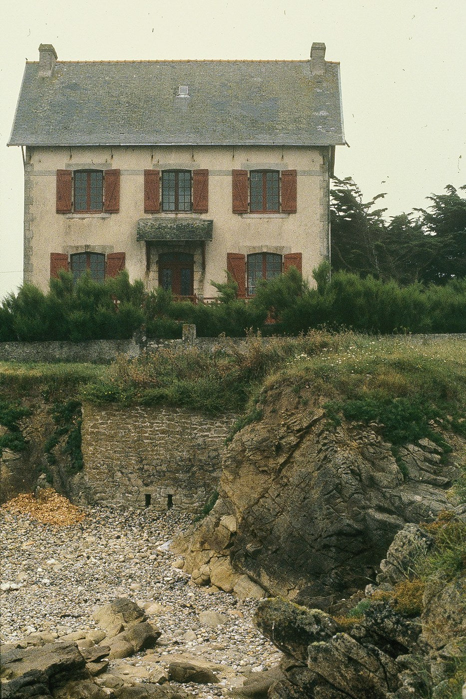 A pale two-story house with red shutters stands above rough coastal ground in Ireland.