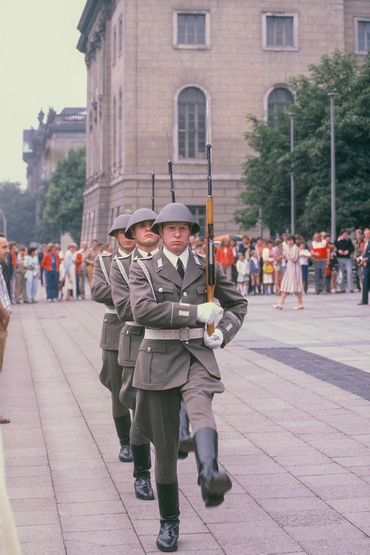 Three uniformed guards march with rifles past spectators across paving beside a building in East Berlin.