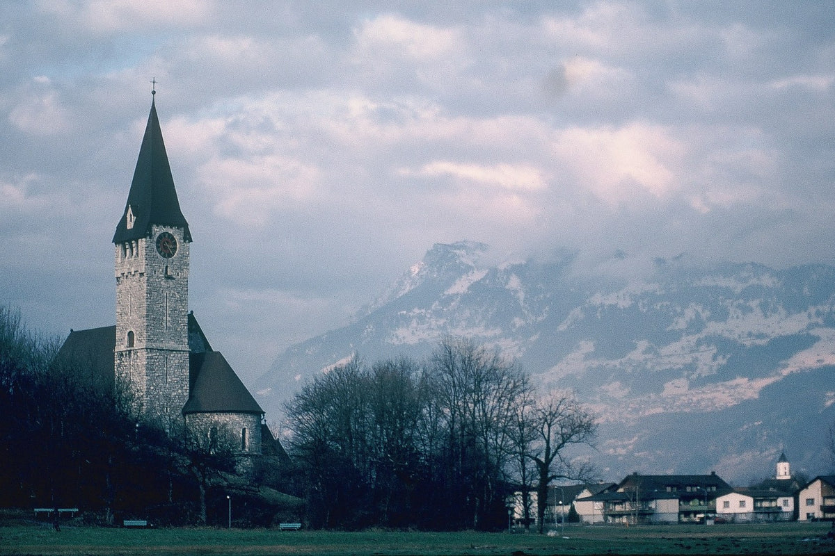 A stone church steeple rises above Bavarian village rooftops with snowy mountains beneath a pastel sky.