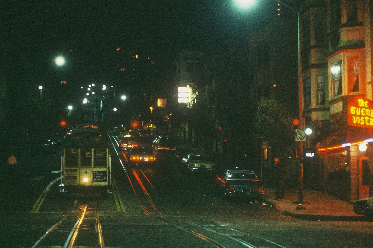 A cable car travels along illuminated tracks at night near San Francisco's Buena Vista sign.