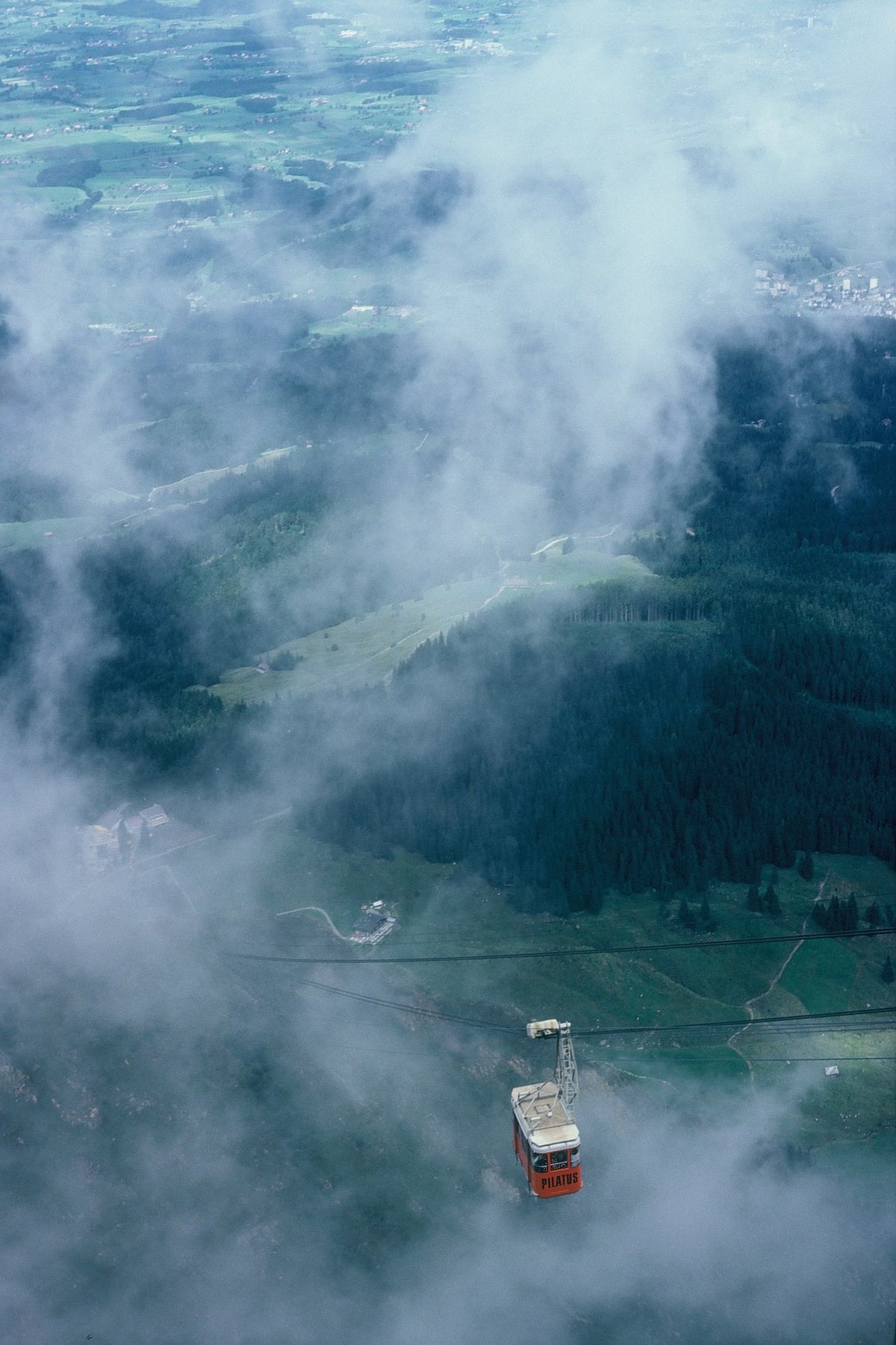 A red cable car hangs above a green Swiss valley partly hidden by clouds, seen from Mount Pilatus.