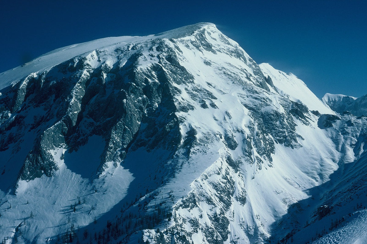 Wind-shaped snow covers a steep mountain summit beneath a deep blue sky.