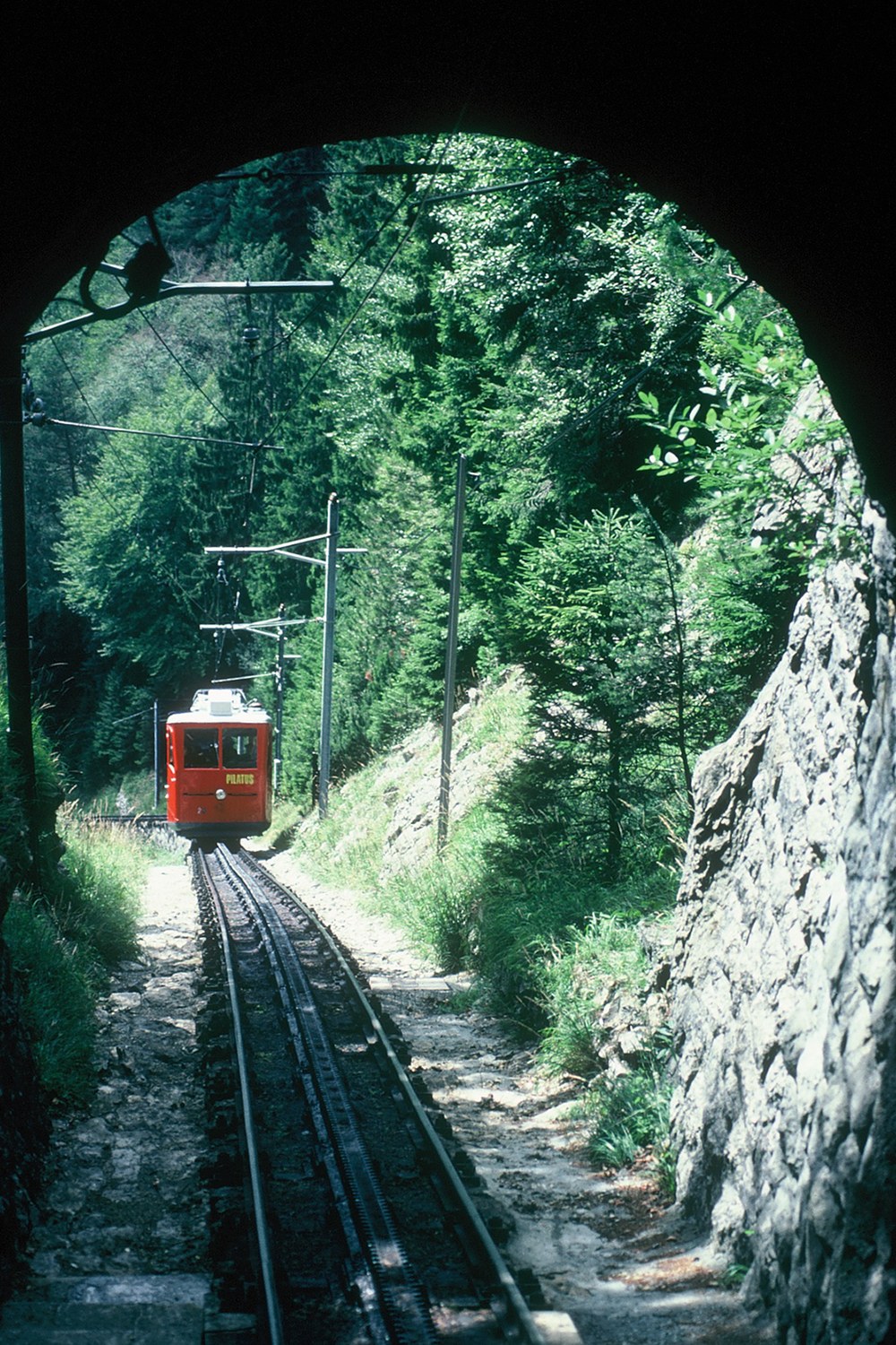 A red railcar approaches on steep track beyond a dark stone tunnel at Mount Pilatus, Switzerland.