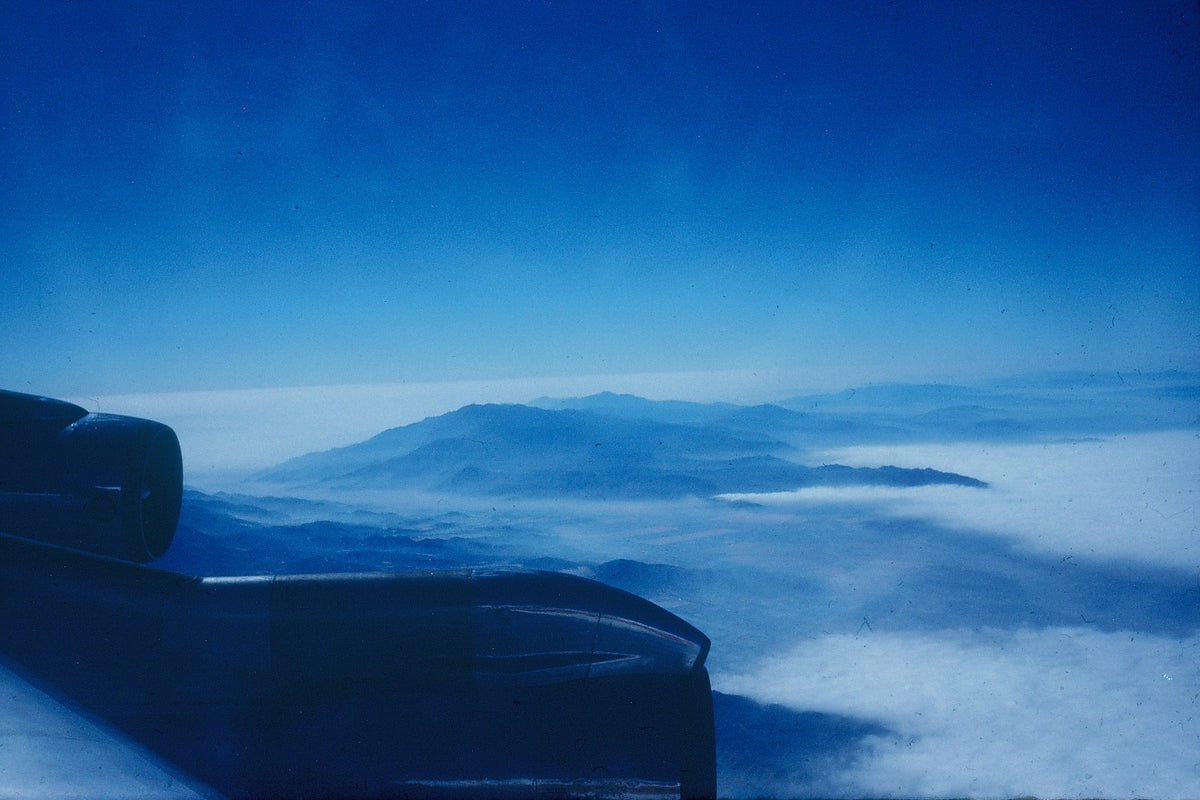 An aircraft engine fills the lower left of a blue aerial view above clouds and distant terrain.