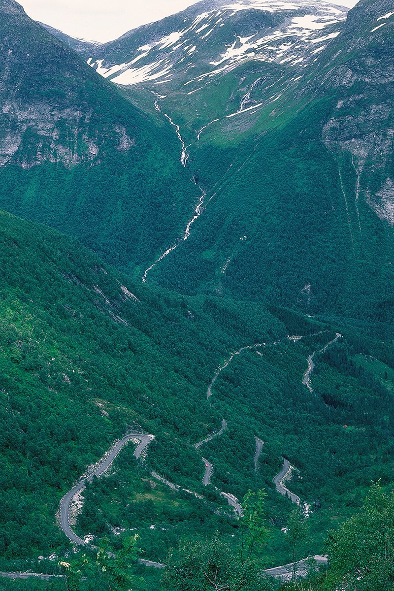 A narrow switchback road winds through a steep green valley in Norway below waterfalls and snowy ridges.