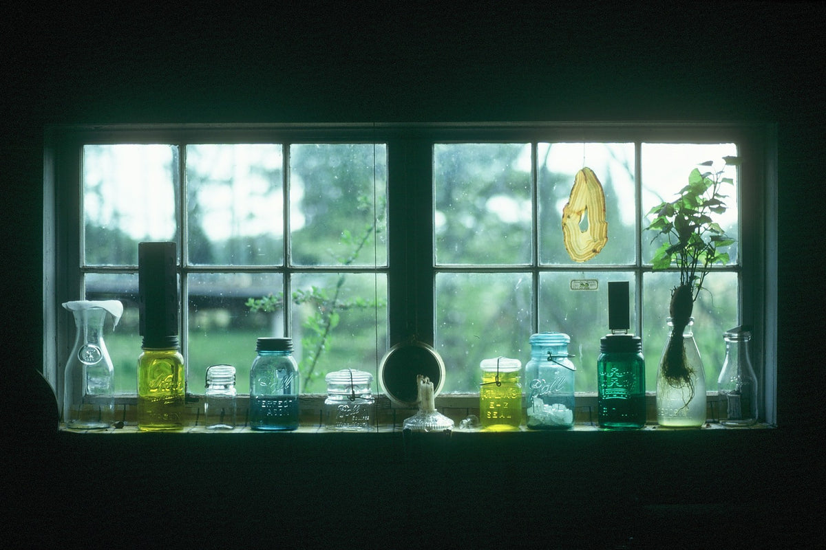 Glass bottles and jars catch green light on a windowsill inside a reconstructed Williamsburg farmhouse.